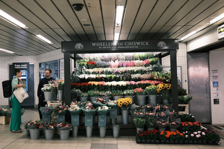 A flower stall named 'Wheeler of Chiswick' located inside a modern underground station, featuring an extensive display of fresh floral arrangements. The arrangement includes a variety of flowers such 