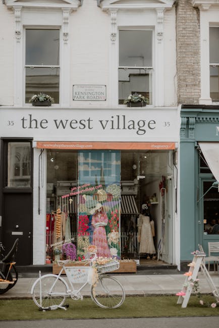 A floral display outside a boutique shop with large windows and white bricks, featuring a colorful array of fresh flowers arranged in a bouquet and displayed on a mannequin dressed in a pink dress. Th