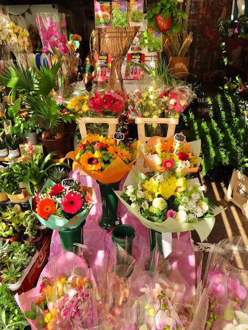 A vibrant display of fresh floral bouquets arranged on a pink table within a florist shop. The bouquets feature a variety of flowers including bright pink, yellow, orange, and white blooms, with some 