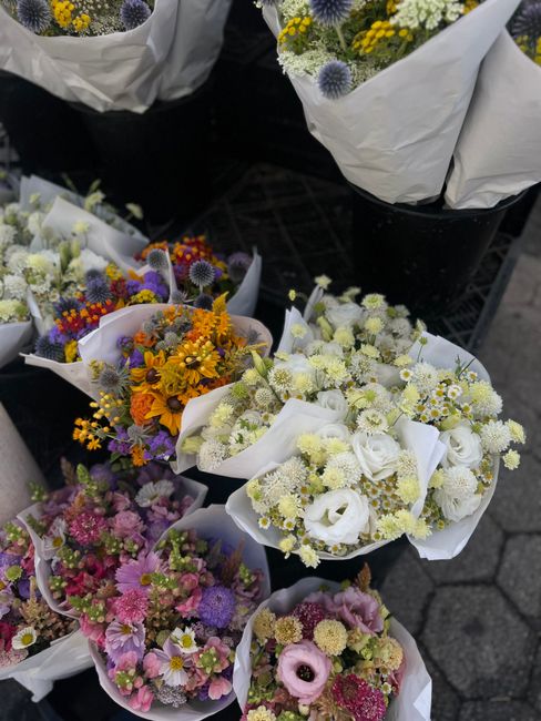 An array of fresh floral bouquets featuring a variety of seasonal flowers arranged with white wrapping paper, displayed outdoors on a black surface. The bouquets include a mix of vibrant colors such a