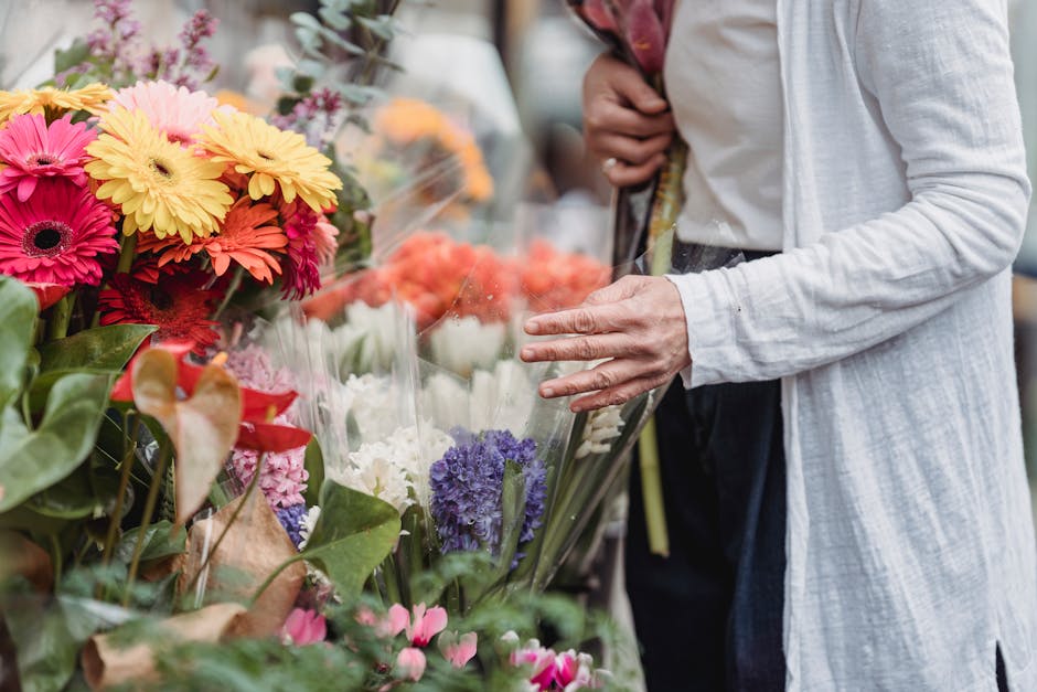 A person wearing a light-colored, long-sleeved shirt is selecting a bouquet of fresh flowers at a floral market, with vibrant blooms such as pink, yellow, purple, and white gerbera daisies, hyacinths,