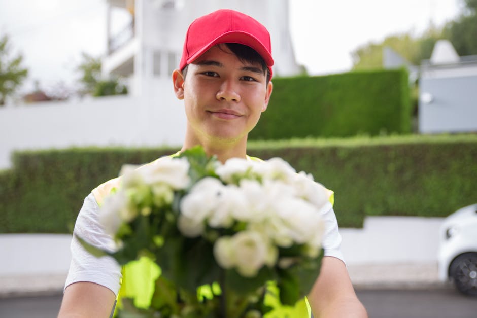 A delivery person wearing a red cap and high-visibility vest is smiling warmly while holding a bouquet of fresh white flowers, including roses and possibly lisianthus, wrapped in clear cellophane. The