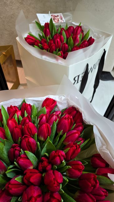 Two bouquets of vibrant red tulips with closed petals and fresh green leaves, arranged in white tissue paper wraps. One bouquet is placed inside a white shopping bag with black lettering, while the ot