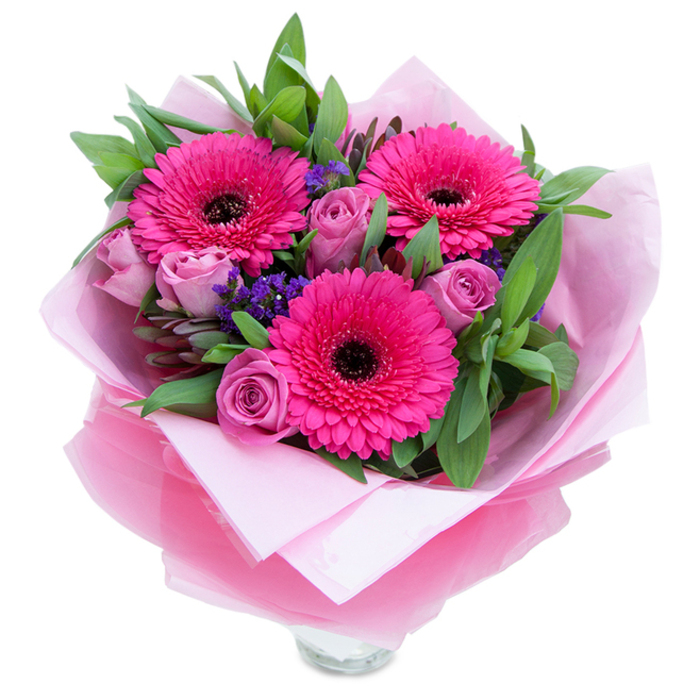 The composition opens with three bold fuchsia Gerbera daisies, their broad, symmetrical petals radiating an energetic pink that immediately draws the eye. Nestled close are roses in a muted lavender-pink or dusty rose shade, their layers of velvety petals still gently cupped as in the first stages of bloom. Between and beneath these larger flowers, sprays of tiny purple statice lend an informal, meadow-like texture that softens the arrangement and adds visual rhythm. A backdrop of rich green foliage - leaves that resemble salal or eucalyptus - anchors the bouquet with a cool, botanical weight and a faintly dewy suggestion.
Careful wrapping transforms the hand-tied stems into a present: an inner sheet of matte pale pink tissue paper provides a soft, supportive base while an outer veil of translucent fuchsia-pink cellophane adds a subtle shimmer and reinforces the monochrome theme. The florist's folds are deliberate and neat, exposing the top rim of a clear glass vase which hints at the freshness and practical display of the flowers. The lighting in the image suggests soft daylight, allowing the gerberas' vivid pigment to sing while revealing the roses' more delicate tonal shifts.
This bouquet feels like a floral embrace - playful yet elegant - suitable for celebrating birthdays, anniversaries, or simply offering encouragement. The scent profile leans to tender rose perfume with a mild green sweetness from the gerberas. Reminiscent of a posy you might pick up on Chiswick High Road or bring home after a stroll through Chiswick House & Gardens, it's crafted to feel intimate, uplifting, and reliably fresh.